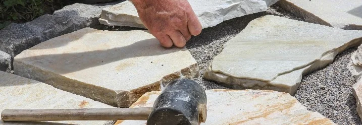 Eine Person verlegt mit einem Gummihammer unregelmäßige Terrassenplatten aus Naturstein.